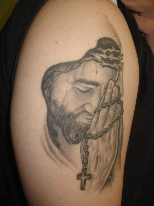 Tatuaje de Jesucristo al que se ha terminado con una especie de capucha que no se ha tatuado y que se enceutnra con las manos en posición de oración y un rosario entre ellas, un brillante tatuaje sin duda alguna