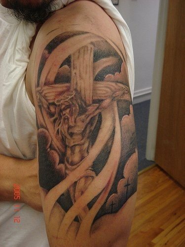 Tatuaje en el brazo de Cristo crucificado sobre un fondo de nubes y viento que dan un espectacular acbado al brazo de este hombre, un tatuaje muy bien pensado y muy bien llevado a cabo